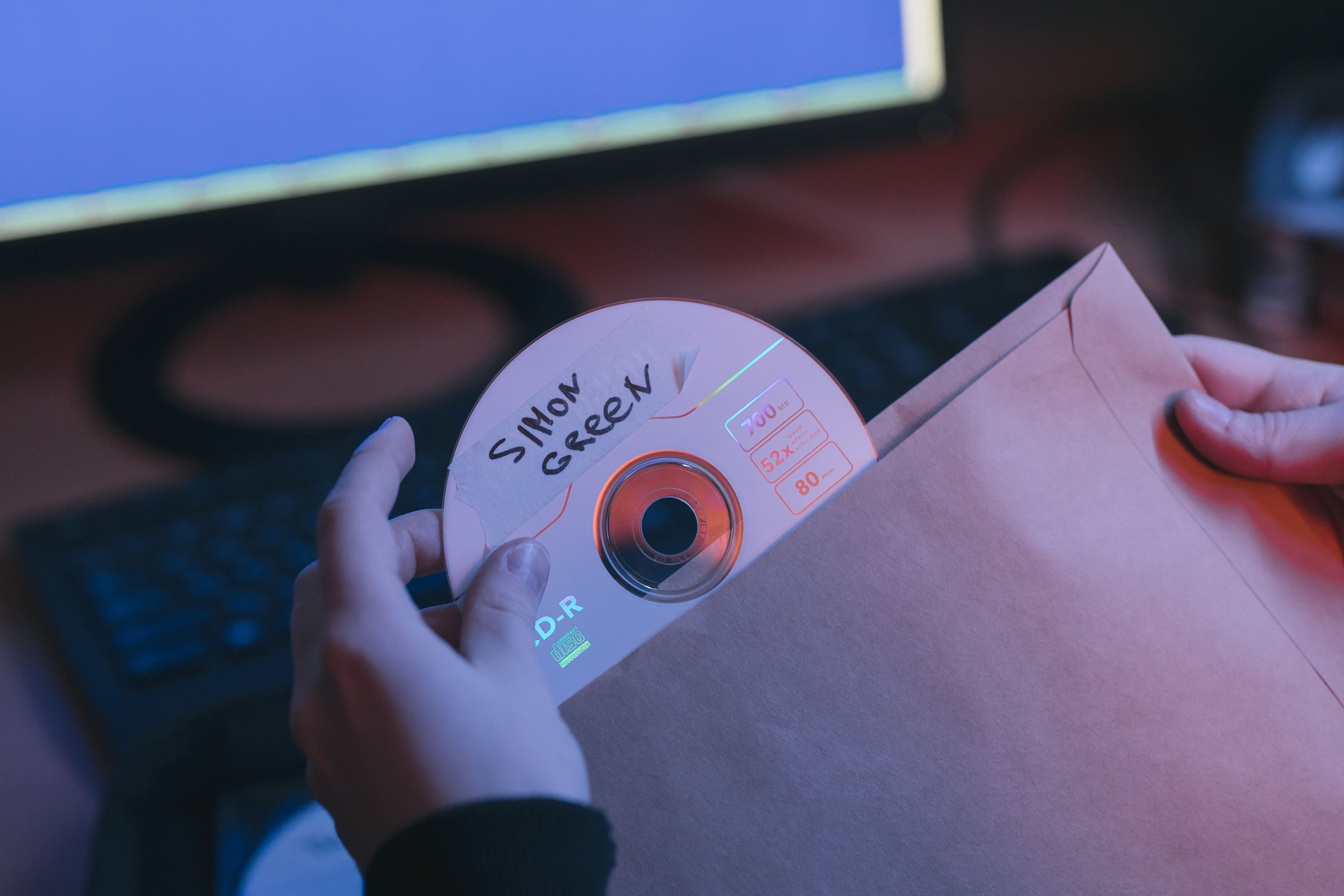 A close-up shot of hands inserting a handwritten CD into an envelope, with a computer in the background.