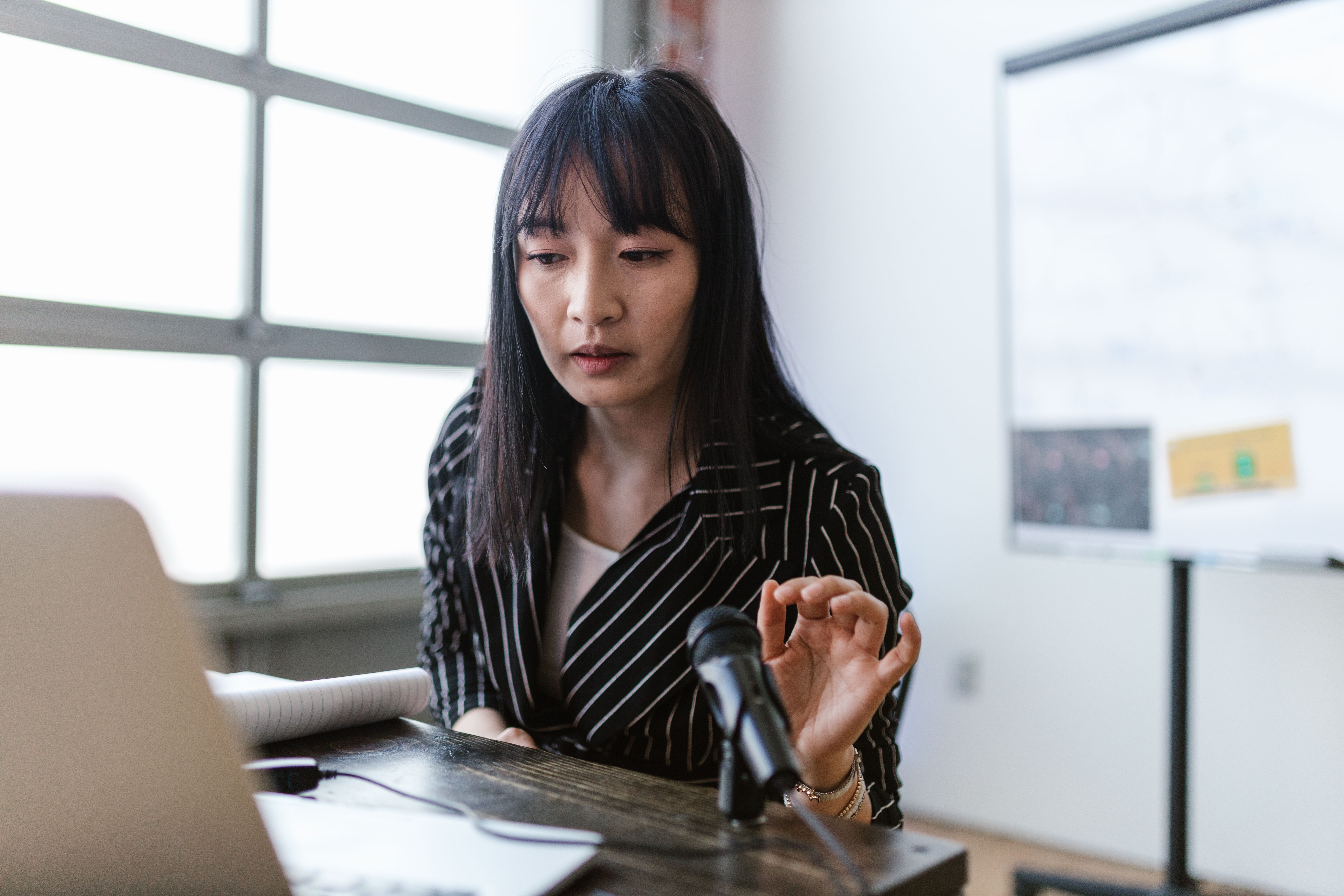 Asian woman in office presenting at a desk with a microphone, wearing striped shirt.