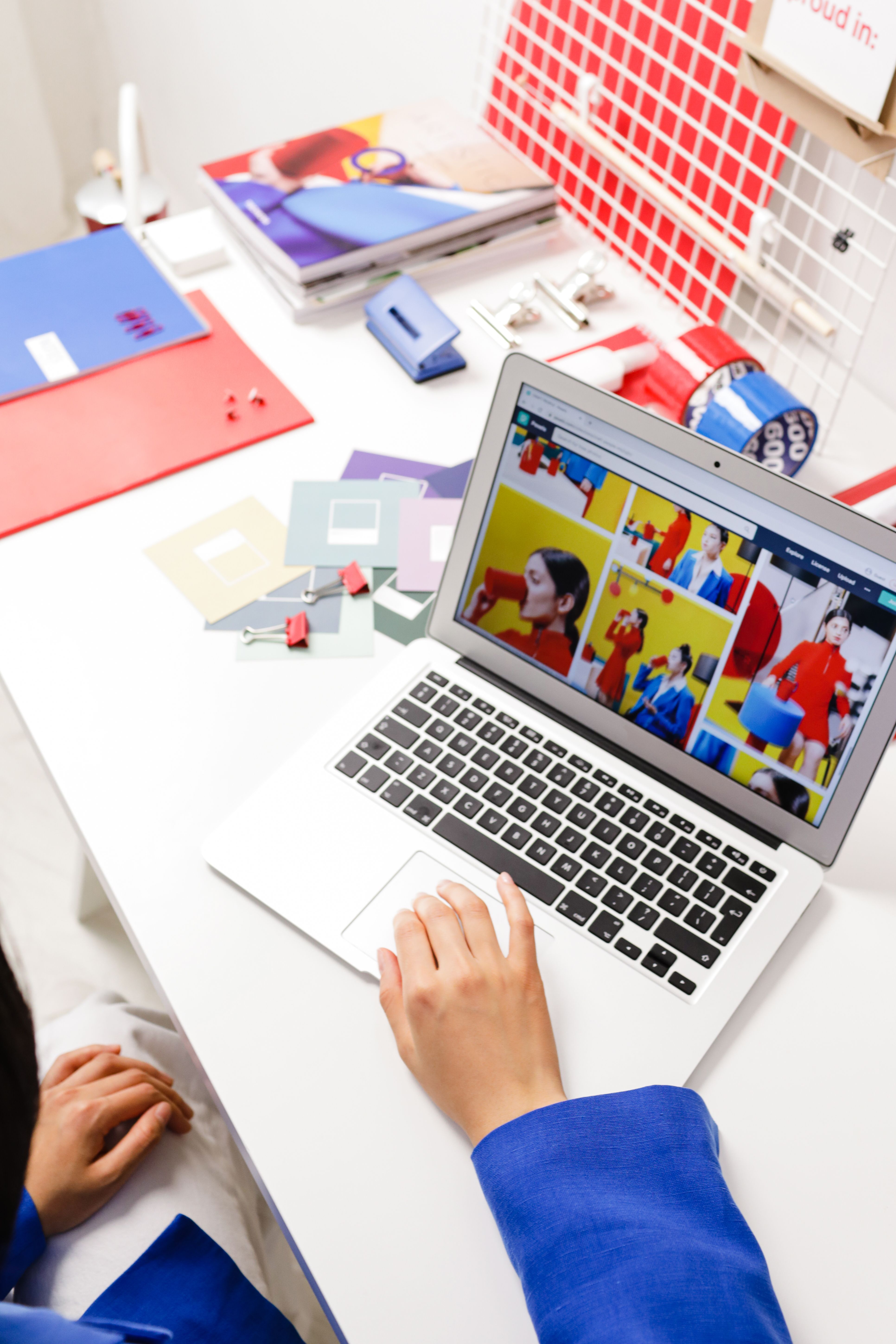 Female in blue jacket using laptop at vibrant desk with notebooks and color samples.